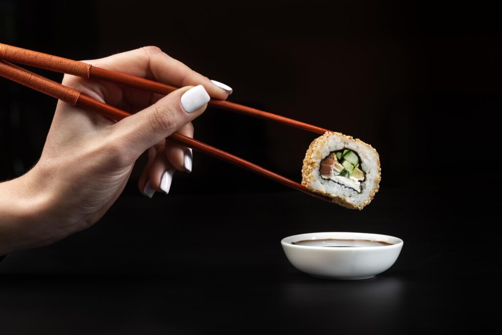 Hand som håller en sushiroll med pinnar över en skål med soja